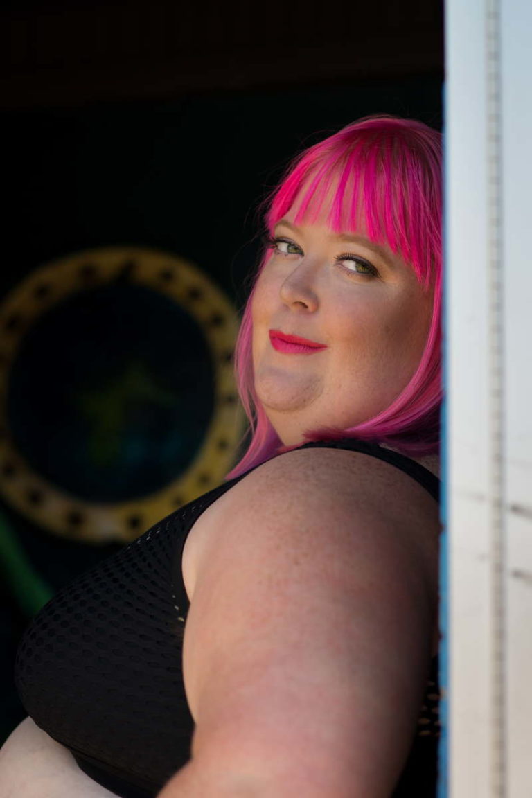 A close-up photo of a fat white person with bright pink hair and lipstick, looking over their shoulder towards the camera. They're wearing a sleeveless black top and leaning against a wall with a nautical-style porthole in the background.