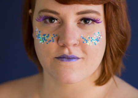 A fat white woman, shown from the bare shoulders up, looks into the camera with a slightly challenging gaze. Her red hair is cut short on one side and jaw-length on the other. She is wearing purple lipstick and false eyelashes, and has large glitter on her cheeks.