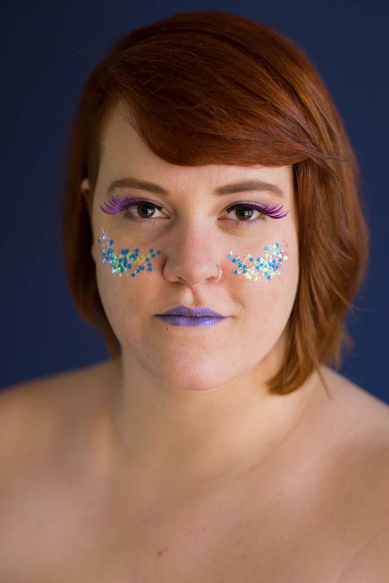 A fat white woman, shown from the bare shoulders up, looks into the camera with a slightly challenging gaze. Her red hair is cut short on one side and jaw-length on the other. She is wearing purple lipstick and false eyelashes, and has large glitter on her cheeks.