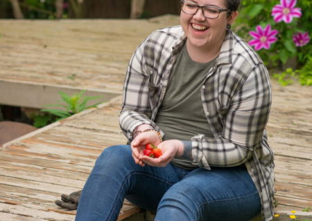 A fat non-binary person with short blue hair sits on a low wooden deck, holding some strawberries, with their feet in a garden bed full of flowers. They're wearing a plaid shirt, jeans and casual shoes.
