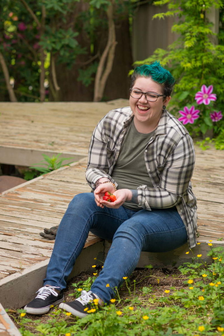 A fat non-binary person with short blue hair sits on a low wooden deck, holding some strawberries, with their feet in a garden bed full of flowers. They're wearing a plaid shirt, jeans and casual shoes.