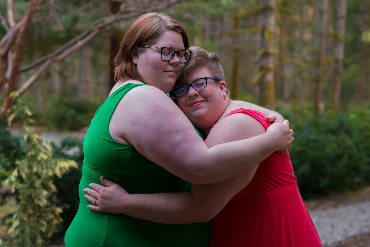 Two fat white women in brightly-colored sleeveless dresses hug on a cloudy day in a park full of trees.