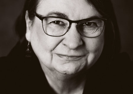 A black-and-white headshot photo of an older woman with facial wrinkles and glasses. Her arms are crossed and she's looking into the camera with a slight smile.
