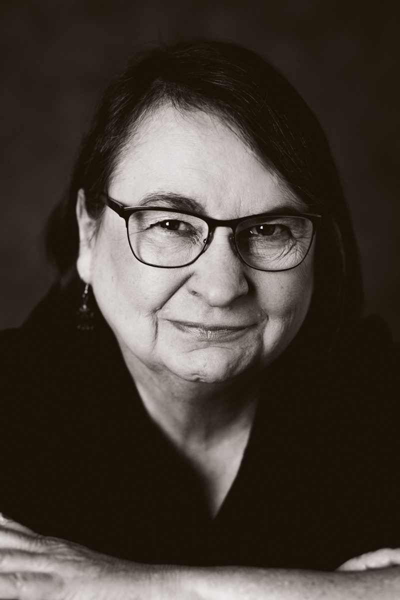 A black-and-white headshot photo of an older woman with facial wrinkles and glasses. Her arms are crossed and she's looking into the camera with a slight smile.