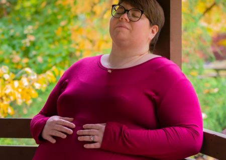 A fat white woman with short brown hair, glasses and a bright pink dress leans back on a wooden bench in a park with fall leaves with a peaceful expression.