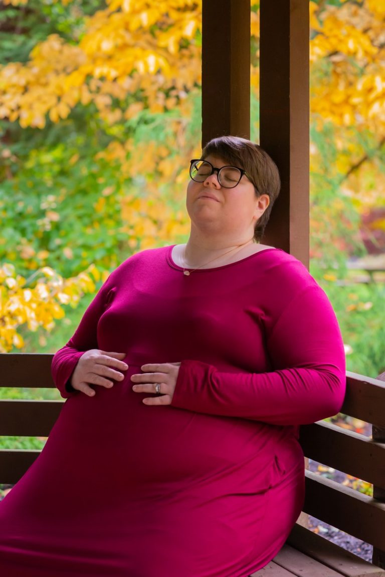 A fat white woman with short brown hair, glasses and a bright pink dress leans back on a wooden bench in a park with fall leaves with a peaceful expression.