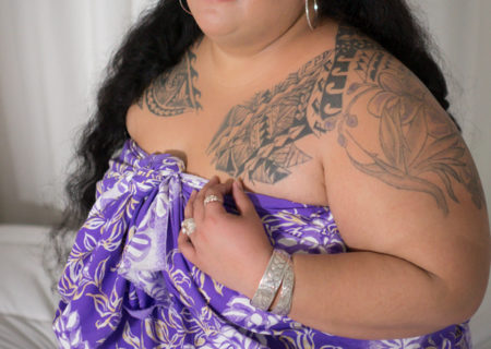 A fat Hawaiian and Tongan woman with brown skin, long black hair and traditional-style tattoos kneels on a bed and looks at the camera with a neutral gaze. She is wearing a purple pareo with white flower designs.