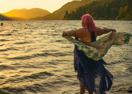 A fat white person with pink hair and a purple dress stands ankle-deep in a mountain lake at sunset, holding a wing-design scarf that floats in the breeze behind them.