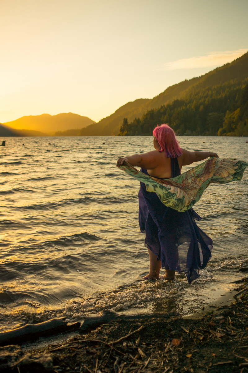 A fat white person with pink hair and a purple dress stands ankle-deep in a mountain lake at sunset, holding a wing-design scarf that floats in the breeze behind them.