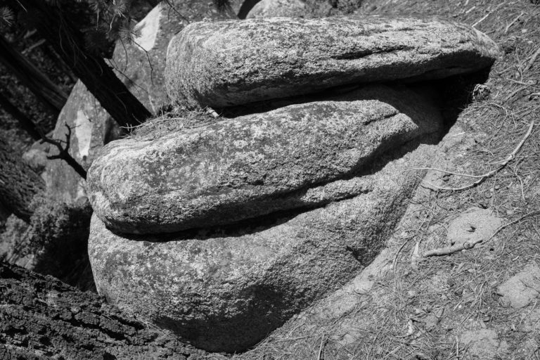 A black-and-white photo of a boulder eroded into three sections sitting on mostly-bare ground. The image is rotated so that the sections of the boulder appear horizontal rather than vertical.