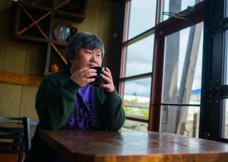 A fat Chinese-American woman with short black and gray hair sits at a coffeehouse table with a mug in her hands, looking out the window. She's wearing a purple Black Lives Matter t-shirt and a dark green cardigan.