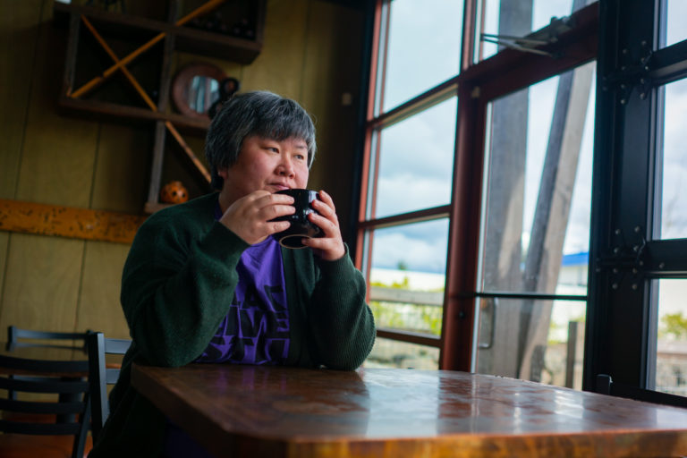 A fat Chinese-American woman with short black and gray hair sits at a coffeehouse table with a mug in her hands, looking out the window. She's wearing a purple Black Lives Matter t-shirt and a dark green cardigan.