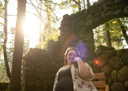 A fat white woman with glasses and long brown hair looks down and to one side, smiling, with one hand to her cheek while sitting on a wooden chair in a ruined stone house in the woods with sun shining through the trees.