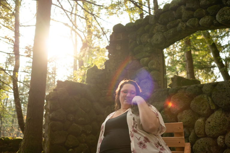 A fat white woman with glasses and long brown hair looks down and to one side, smiling, with one hand to her cheek while sitting on a wooden chair in a ruined stone house in the woods with sun shining through the trees.