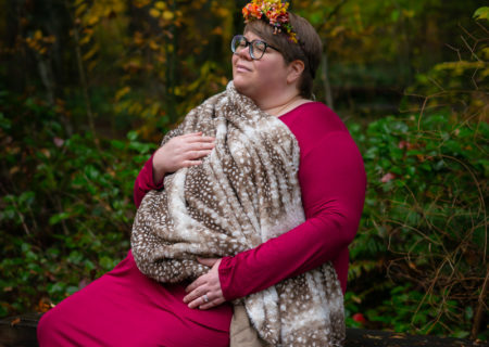 A fat pregnant woman in a floral headband, glasses and a long pink dress holds a spotted faux fur blanket over her torso. She's sitting on a bench in the woods and looking to one side with a neutral expression.