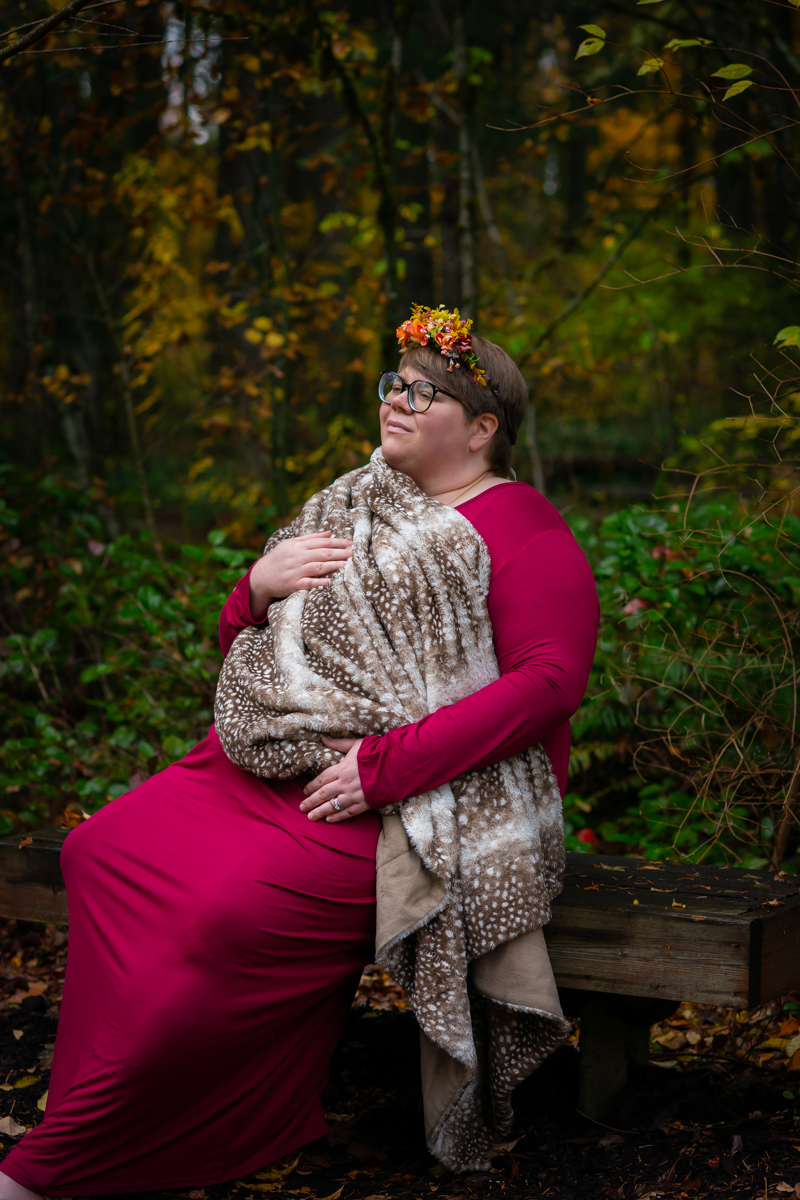 A fat pregnant woman in a floral headband, glasses and a long pink dress holds a spotted faux fur blanket over her torso. She's sitting on a bench in the woods and looking to one side with a neutral expression.