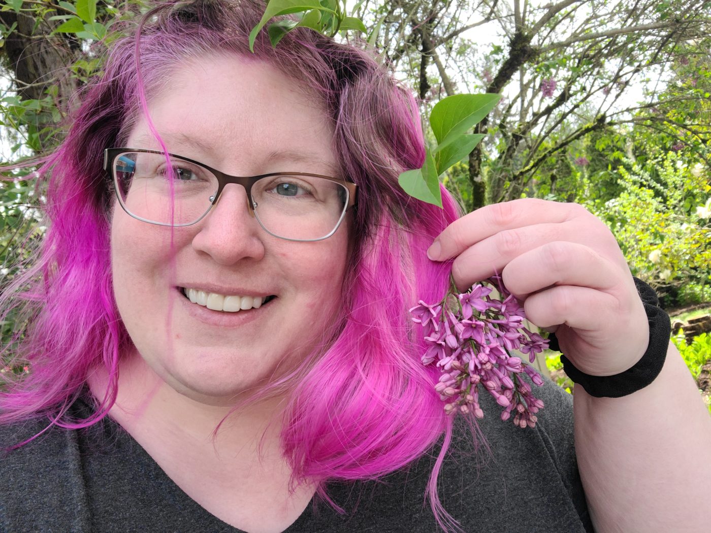 Lindley, a fat white woman with glasses and a gray v-neck top, smiles and holds a flowering lilac branch close to her purple hair.