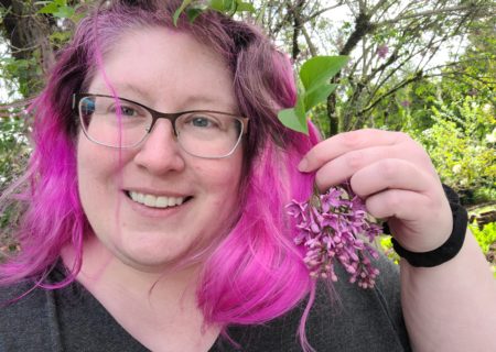 Lindley, a fat white woman with glasses and a gray v-neck top, smiles and holds a flowering lilac branch close to her purple hair.