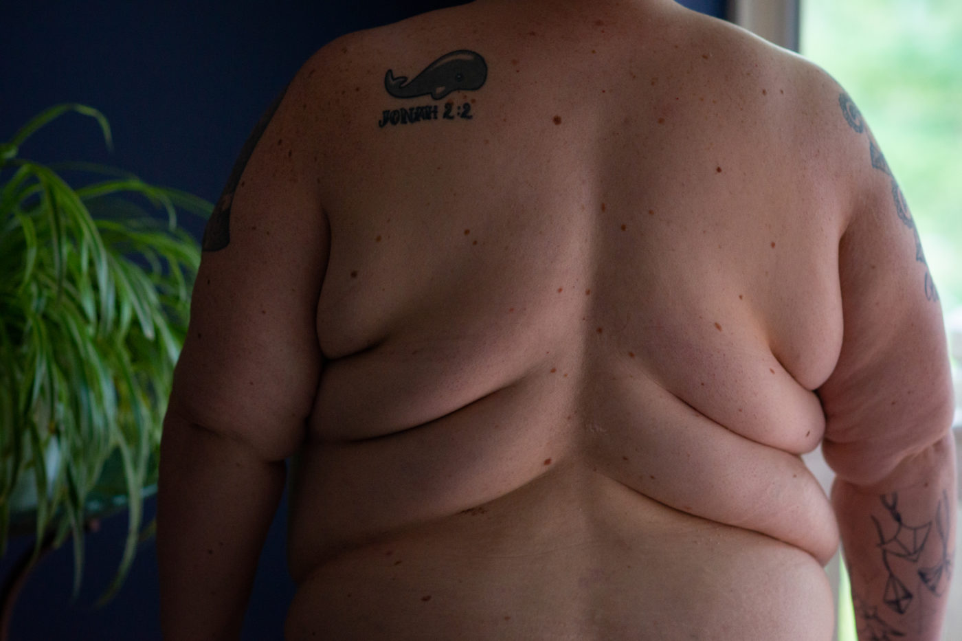 A fat white person's back, back rolls and arms seen in a dark room lit by a window, with a large plant on their other side. On their back is a tattoo of a whale with a Bible verse reference.