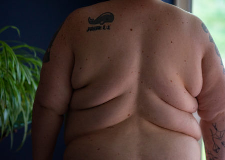 A fat white person's back, back rolls and arms seen in a dark room lit by a window, with a large plant on their other side. On their back is a tattoo of a whale with a Bible verse reference.