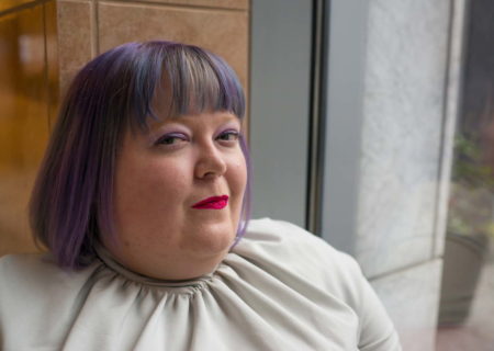 A fat white non-binary person in a gray top with a tight collar sits on a window ledge inside a building with salmon-colored stone tile walls. Their hair is purple. They're wearing eyeshadow and lipstick, and are looking at the camera with a mischievous expression.