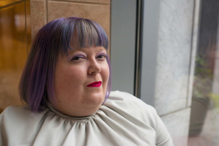 A fat white non-binary person in a gray top with a tight collar sits on a window ledge inside a building with salmon-colored stone tile walls. Their hair is purple. They're wearing eyeshadow and lipstick, and are looking at the camera with a mischievous expression.