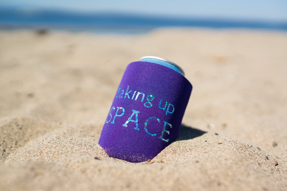 A purple drink koozie with the words Taking Up Space sitting on a sandy beach.