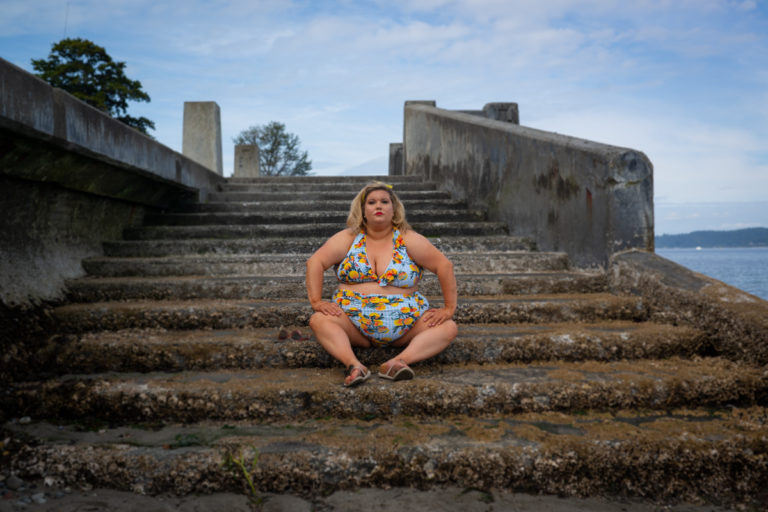 A fat white woman with blonde hair held back by sunglasses wears a blue bikini with an orange pattern. She's sitting on a set of seaweedy concrete water stairs and her body language and expression are serious and challenging.