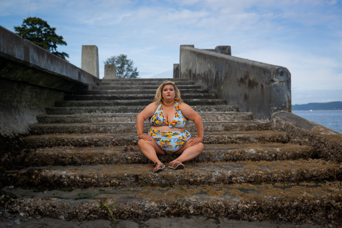A fat white woman with blonde hair held back by sunglasses wears a blue bikini with an orange pattern. She's sitting on a set of seaweedy concrete water stairs and her body language and expression are serious and challenging.