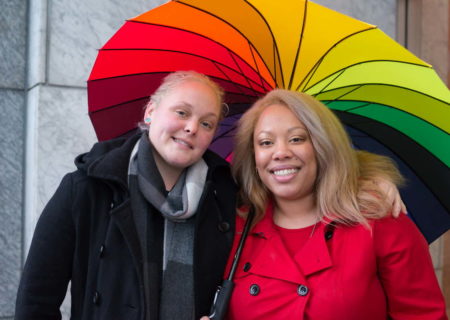 A lesbian couple stands outside a stone building wearing winter coats and smiling under a rainbow umbrella. One is a blonde white woman and the other is a light-skinned Black woman.