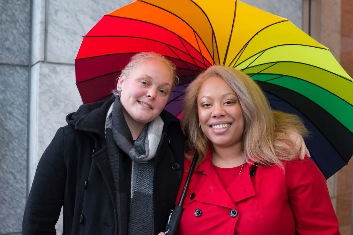 A lesbian couple stands outside a stone building wearing winter coats and smiling under a rainbow umbrella. One is a blonde white woman and the other is a light-skinned Black woman.