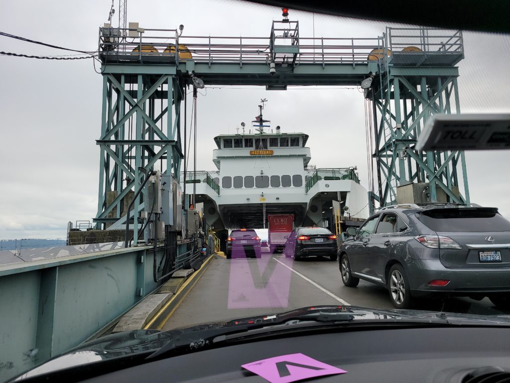 A through-the-windshield photo of driving onto a large ferry beside other cars. In the foreground is a metal structure like a bridge over the road.