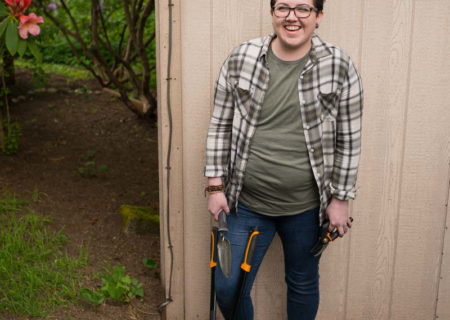 In this free plus-size stock photo, an agender person with short blue hair, a plaid top over a t-shirt and blue jeans holds hedge clippers and stands in front of a wooden shed.