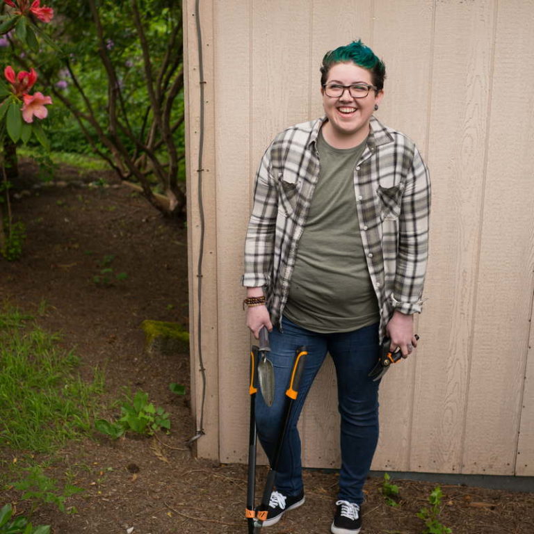 In this free plus-size stock photo, an agender person with short blue hair, a plaid top over a t-shirt and blue jeans holds hedge clippers and stands in front of a wooden shed.