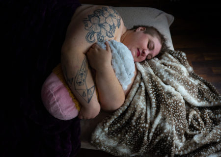 A fat white person with tattoos and short hair lies with eyes closed and a peaceful expression on a brown and white spotted blanket and holds a large stuffed coffee cup with both arms. They're at a Seattle body-positive boudoir session.