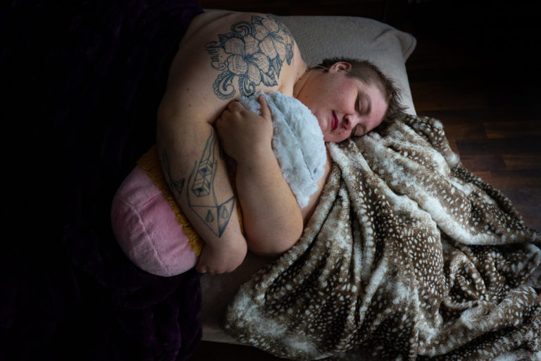 A fat white person with tattoos and short hair lies with eyes closed and a peaceful expression on a brown and white spotted blanket and holds a large stuffed coffee cup with both arms. They're at a Seattle body-positive boudoir session.