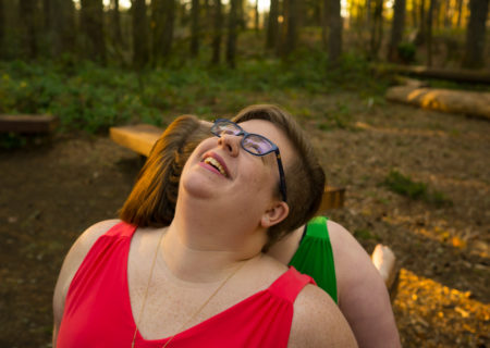 Two fat women are shown from the shoulders up sitting back to back on a bench in the forest, with heads thrown back laughing. The woman whose face is shown has short brown hair and glasses, and both are wearing bright sleeveless dresses. They are at a body-positive portrait photography session in Seattle, WA.