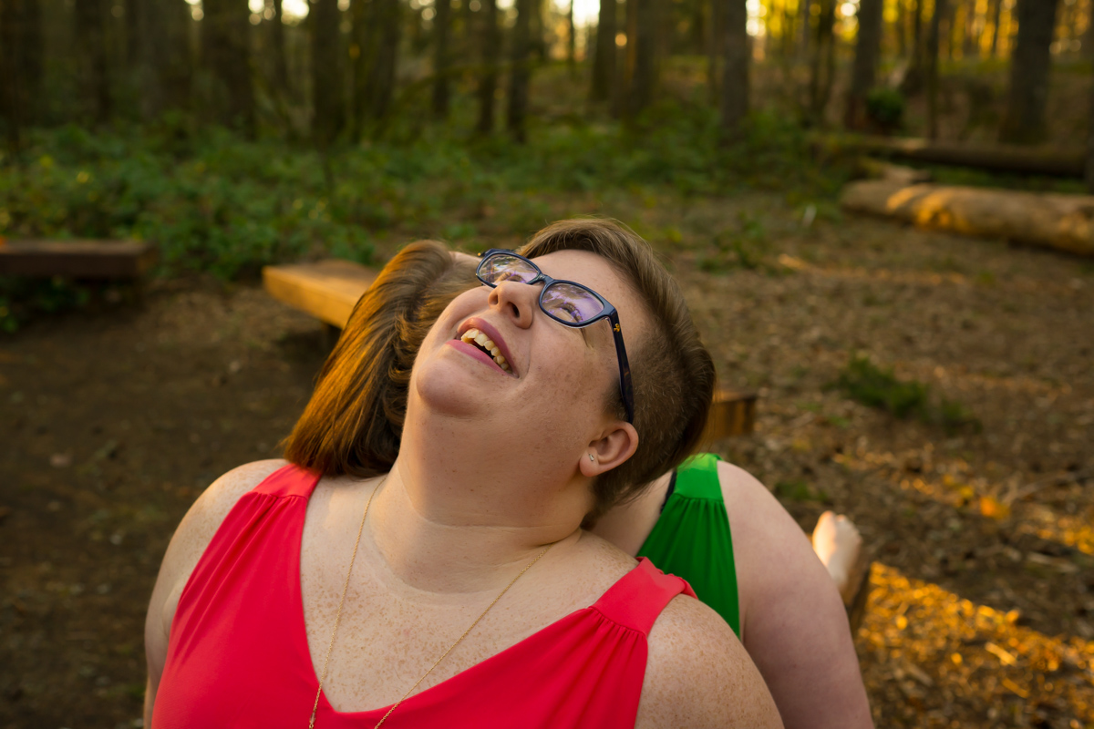 Two fat women are shown from the shoulders up sitting back to back on a bench in the forest, with heads thrown back laughing. The woman whose face is shown has short brown hair and glasses, and both are wearing bright sleeveless dresses. They are at a body-positive portrait photography session in Seattle, WA.