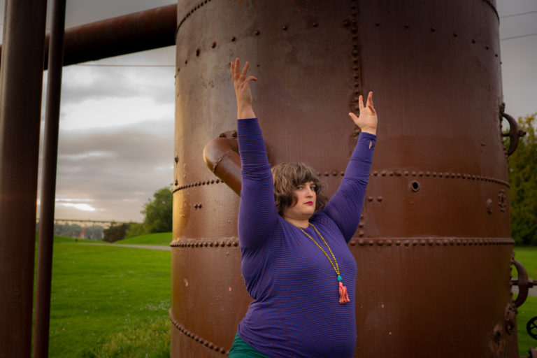 A fat woman with pale skin and curly brown hair, wearing a long-sleeved purple shirt and long mala necklace, raises her arms in a yoga pose in front of a massive brown pipe left over from an old gasworks operation in a city park.