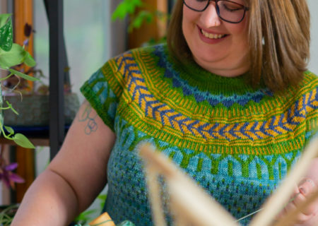 A fat white woman with shoulder-length brown hair and glasses smiles down at yarn she is winding on a wooden device into a spool. She's wearing an intricate handmade sweater at her Seattle body-positive small business portrait photo shoot.