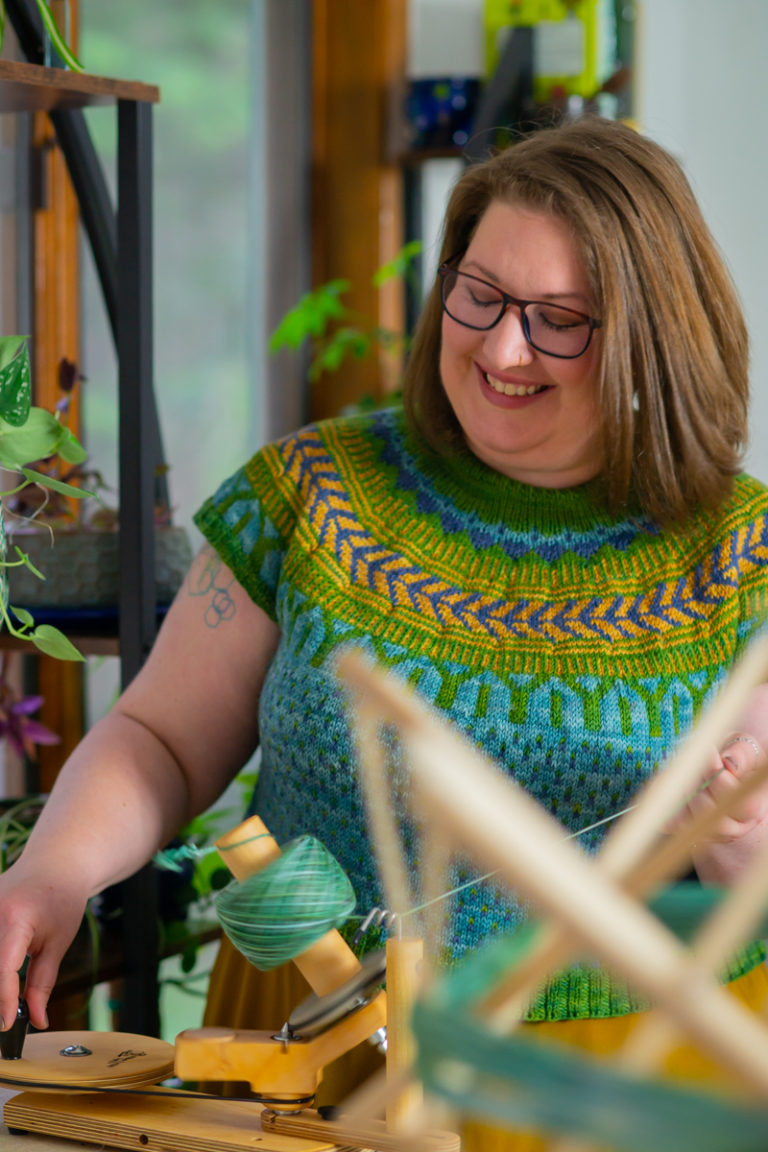 A fat white woman with shoulder-length brown hair and glasses smiles down at yarn she is winding on a wooden device into a spool. She's wearing an intricate handmade sweater at her Seattle body-positive small business portrait photo shoot.