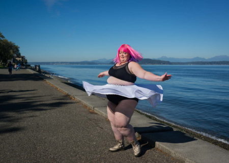 A fat white person with bright pink hair twirls on a walkway in a waterfront park on a sunny day. They're wearing a black bra-like crop top and a white skirt that flares out around them.