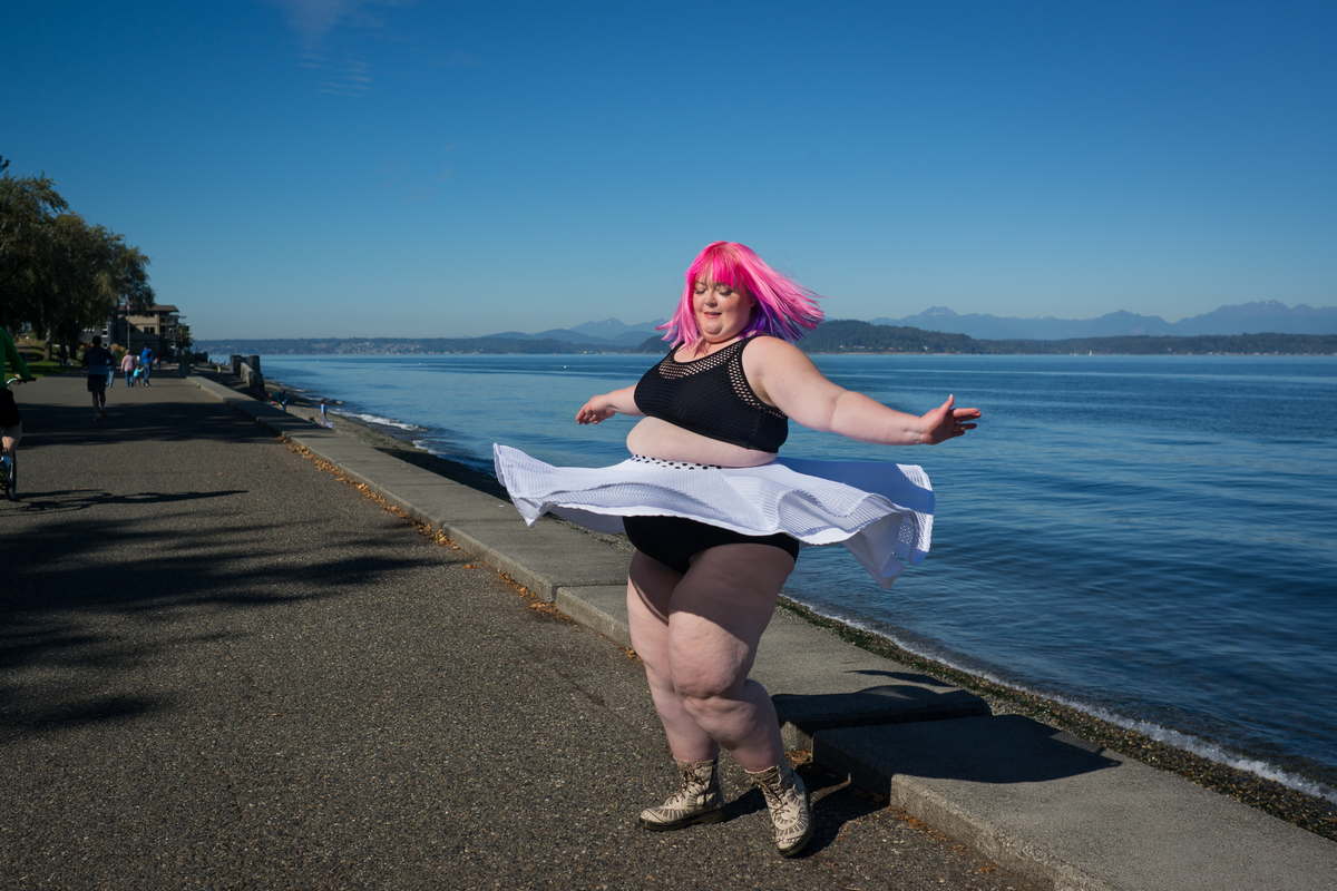 A fat white person with bright pink hair twirls on a walkway in a waterfront park on a sunny day. They're wearing a black bra-like crop top and a white skirt that flares out around them.