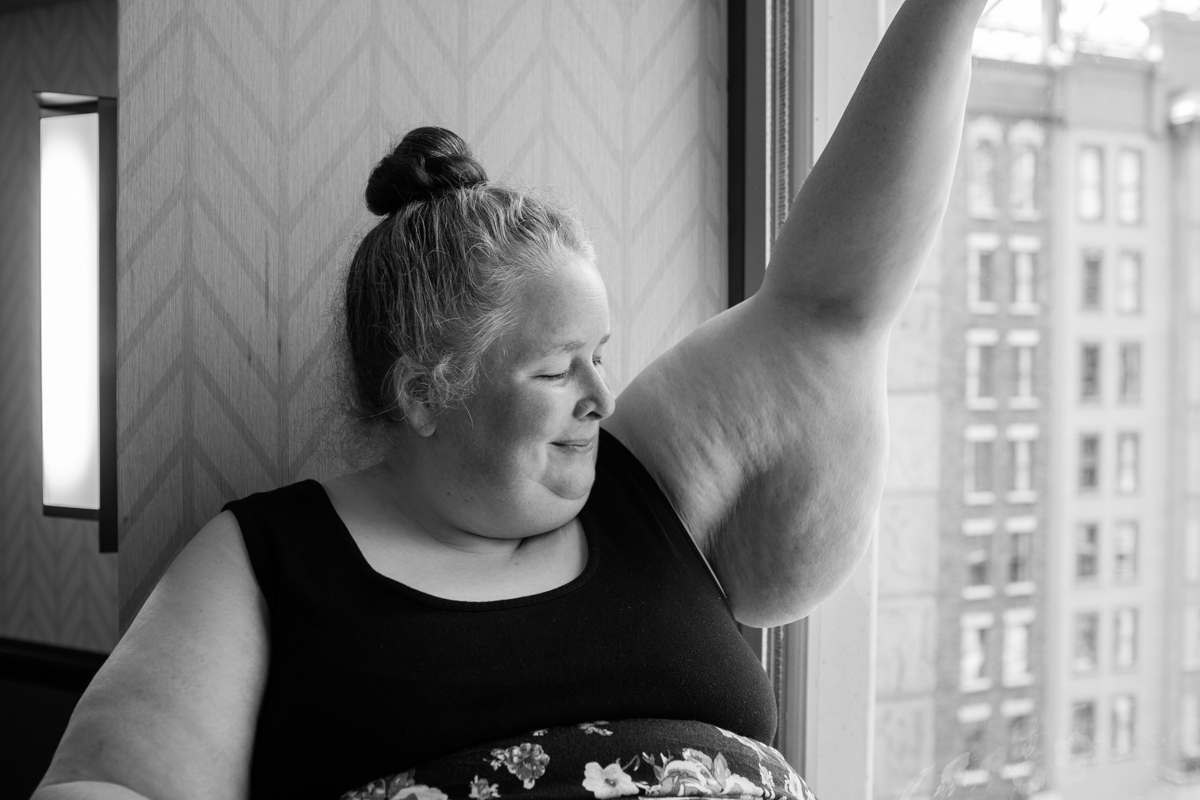 A fat white woman wearing a sleeveless dress and with her hair pulled up into a topknot sits in a window seat, shown from the waist up. She is smiling over at her raised bare arm, which has a large, soft "wing" of skin.
