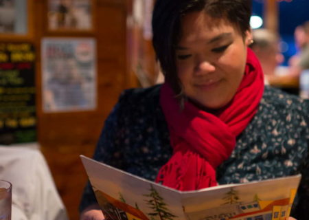 A plus-size woman with Southeast Asian heritage looks at a menu in a rustic seafood restaurant. She has short black hair and is wearing a blue shirt and red scarf.