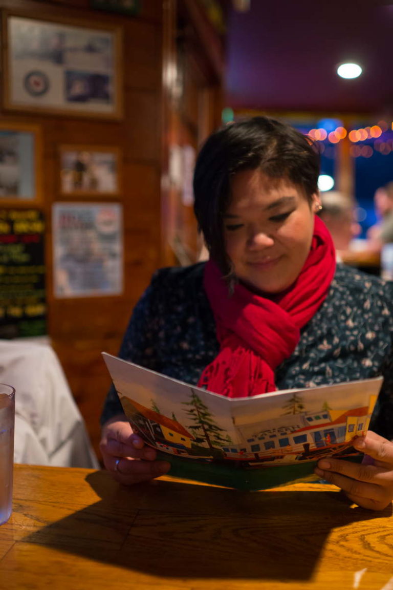 A plus-size woman with Southeast Asian heritage looks at a menu in a rustic seafood restaurant. She has short black hair and is wearing a blue shirt and red scarf.