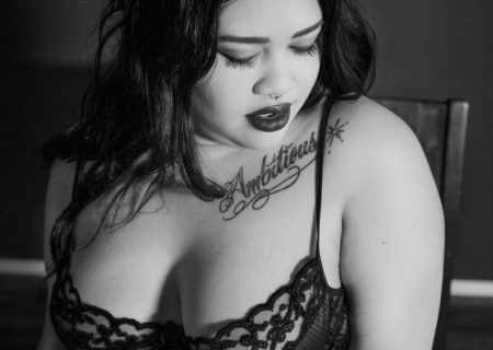A black and white image of a young woman with long black hair, light brown skin, heavy lipstick, a tattoo that says Ambitious, and a lace bra. She's sitting in a chair and looking down and away with a sultry expression at a body positive boudoir photo session in Seattle, WA.