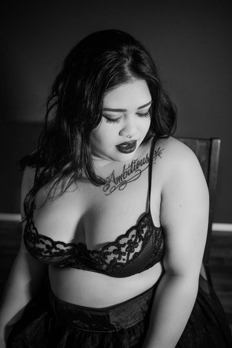 A black and white image of a young woman with long black hair, light brown skin, heavy lipstick, a tattoo that says Ambitious, and a lace bra. She's sitting in a chair and looking down and away with a sultry expression at a body positive boudoir photo session in Seattle, WA.