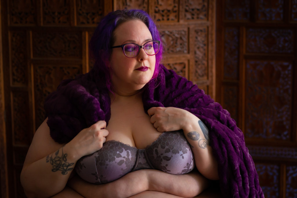 A fat woman with purple hair and matching glasses poses in lingerie with a purple blanket over her shoulders and fat rolls showing
