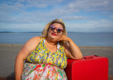 A fat white woman with shoulder-length blonde hair, sunglasses, red lipstick and a sleeveless sundress leans on a red vintage suitcase in front of a body of water in the sun. She is at a Seattle-area body positive portrait photography session.